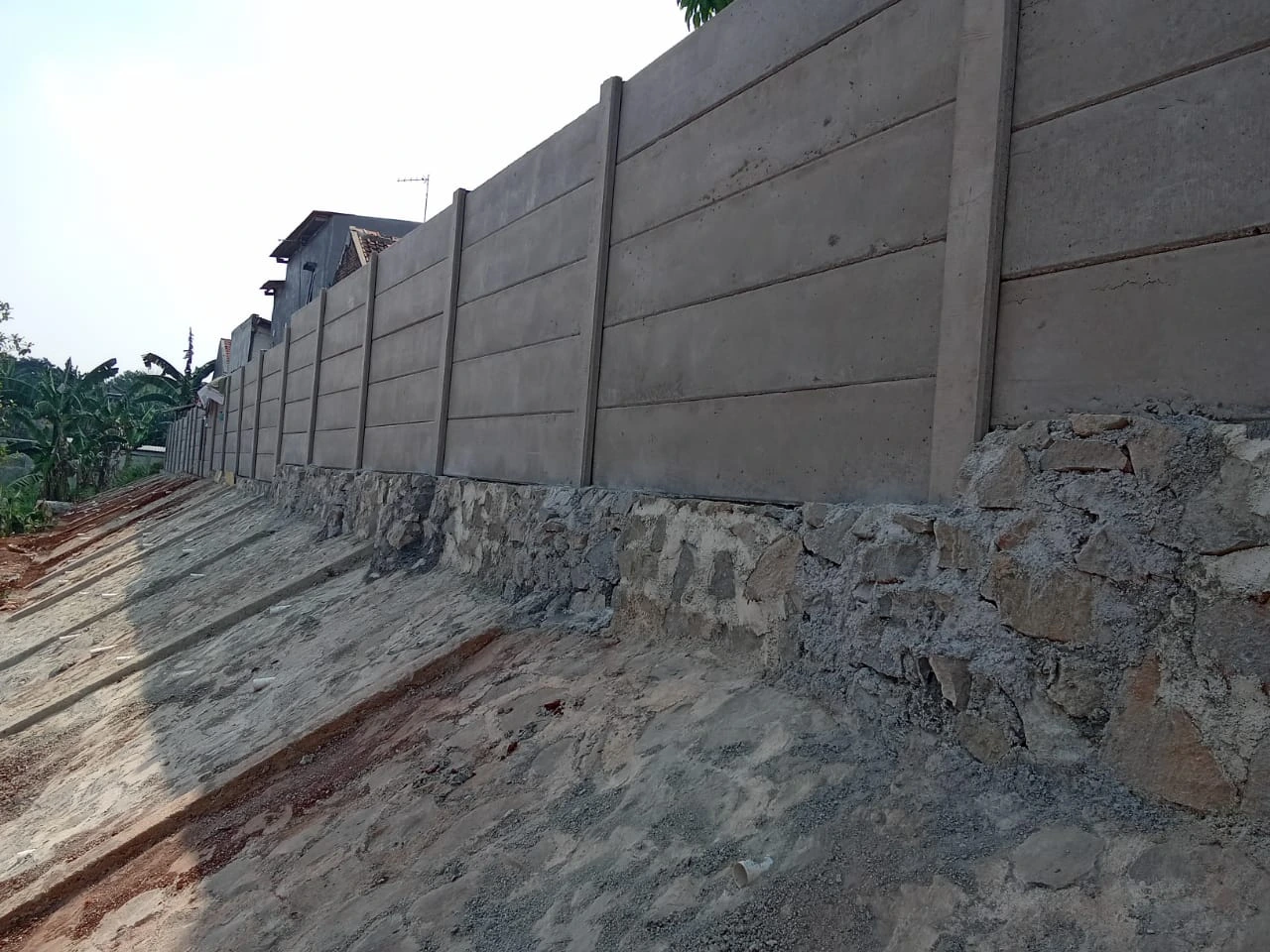 Pagar panel beton berdiri di atas pondasi batu kali pada area tanah miring