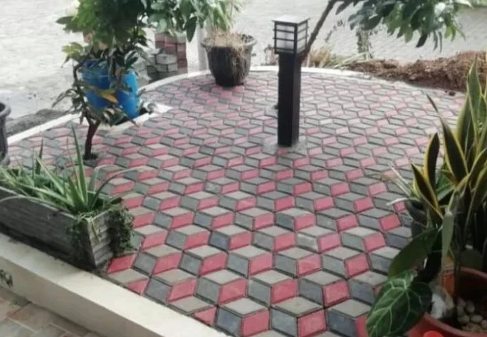 Paving block motif kubus 3D warna merah, abu-abu, dan hitam yang terpasang di area taman rumah dengan lampu taman di tengah.