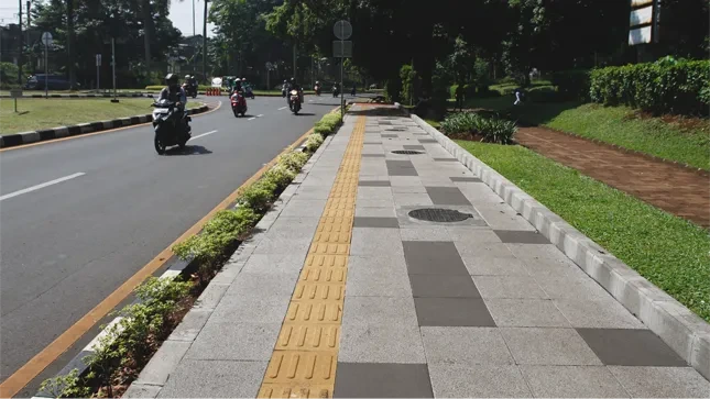 Trotoar dengan kanstin beton di tepi jalan kota, dilengkapi jalur pemandu kuning untuk pejalan kaki dan taman hijau di sisi jalan.