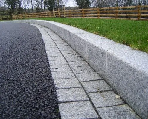Kanstin beton warna abu-abu pada tepi jalan dengan paving block sebagai jalur drainase dan rumput di sisi trotoar.