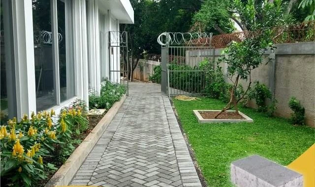 Jalur pejalan kaki dengan paving block dan kanstin beton di halaman rumah modern, dilengkapi taman hijau dan tanaman hias.