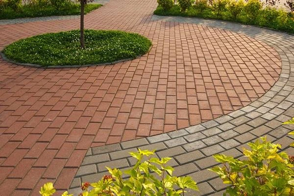 Area taman dengan paving block tersusun rapi menggunakan kombinasi warna merah dan abu-abu membentuk pola melengkung.