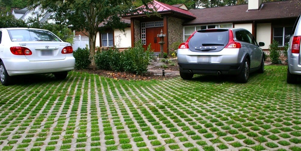Area parkir rumah menggunakan paving block rumput dengan pola grid, di mana beberapa mobil sedang terparkir di atasnya.