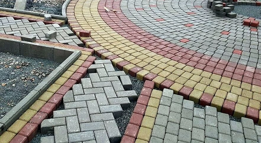 Pemasangan paving block berbagai warna dengan pola melingkar dan bata yang sedang dikerjakan di area halaman.