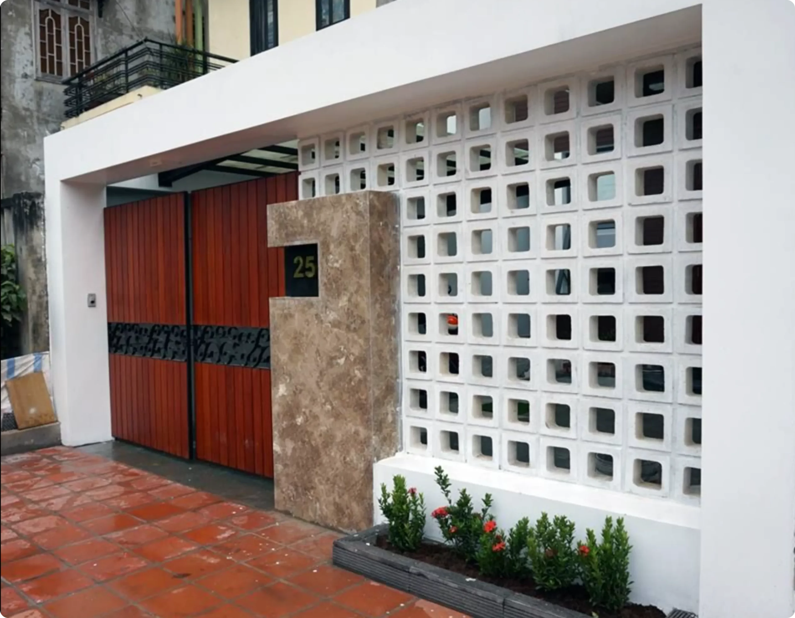 Tampak depan pagar rumah modern dengan dinding roster beton berbentuk kotak-kotak, pintu gerbang kayu merah, dan tanaman hias di bagian bawah.
